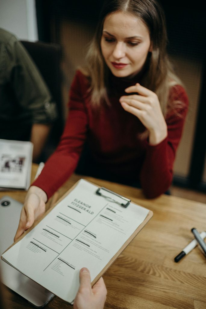 Comment se démarquer des autres candidats et décrocher l’emploi de vos rêves ? Woman sitting at a desk holding a clipboard and reviewing a resume during a job interview.