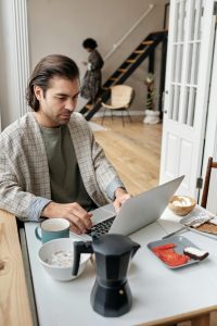 Comment transformer un refus d’embauche en opportunité ? Man working remotely at breakfast table with coffee and laptop in cozy apartment setting.