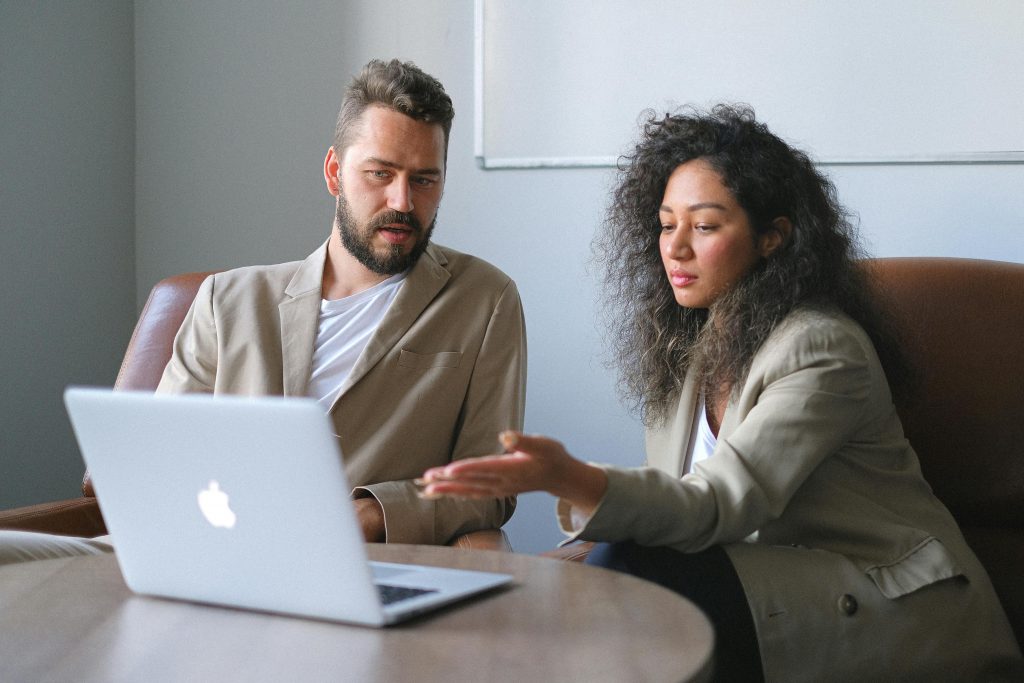 Serious colleagues in stylish outfits sitting at table and using laptop while discussing new project in office