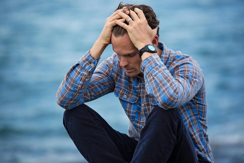 Comment transformer un refus d’embauche en opportunité ? A man in a plaid shirt sits by the water looking distressed, symbolizing stress.
