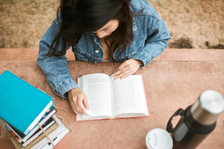 Overhead view of a woman reading a book outdoors on a sunny day, wearing a denim jacket.