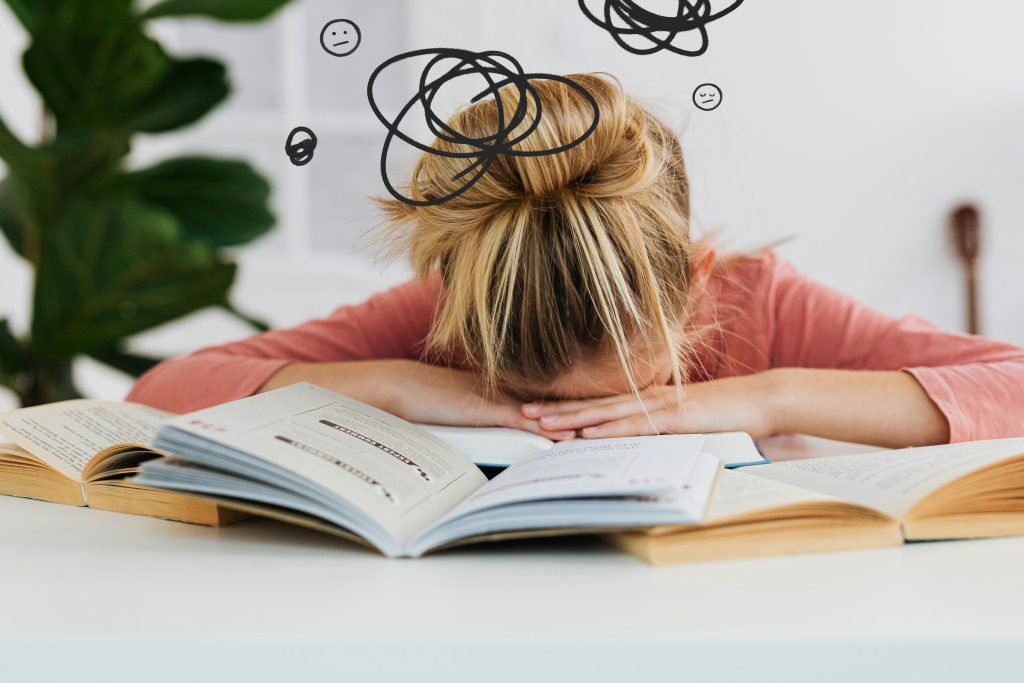 Pourquoi remettons-nous toujours à demain et comment arrêter ? Young woman asleep over books at desk, conveying stress and mental overload.