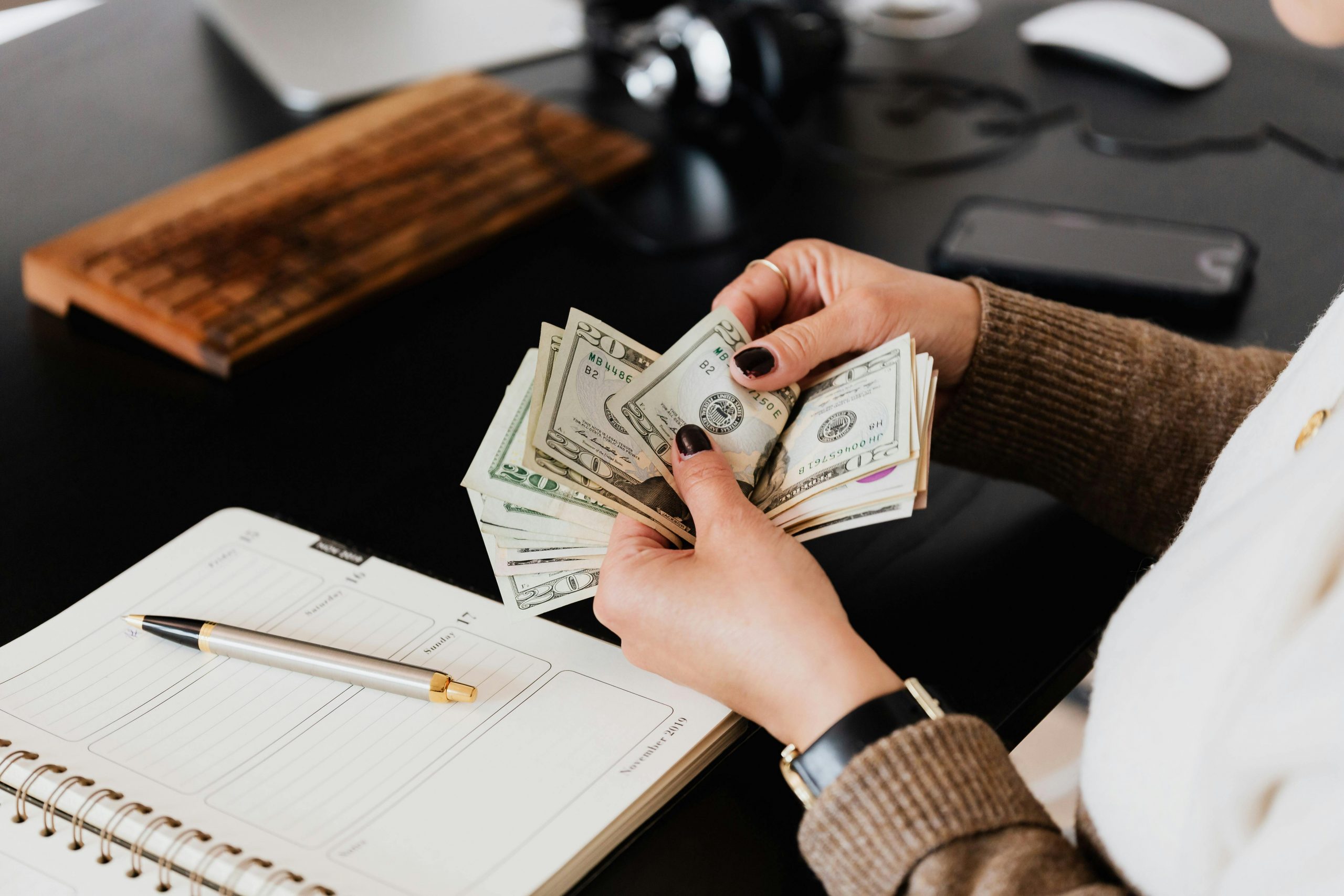 Fonds d’urgence : pourquoi et comment le constituer (guide débutant et intermédiaire) Unrecognizable elegant female in sweater counting dollar bills while sitting at wooden table with planner and pen
