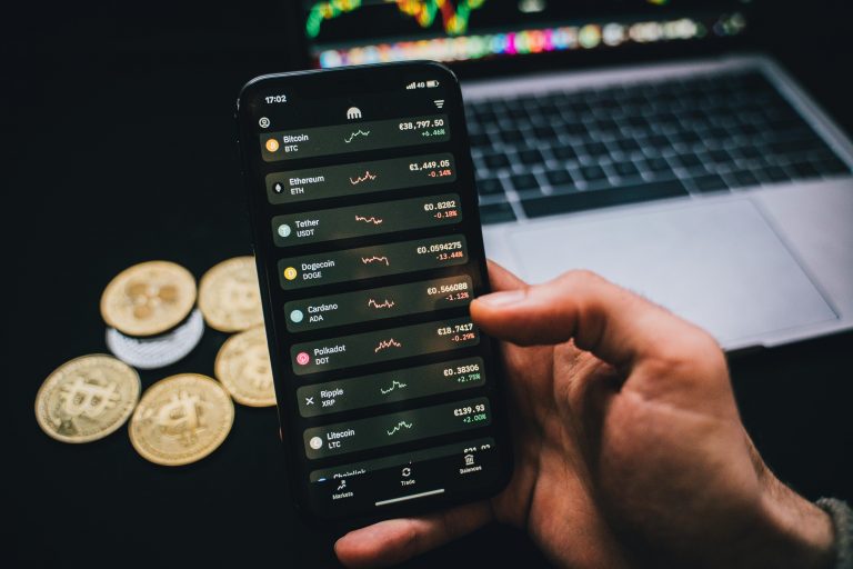 A person checks cryptocurrency values on a smartphone with a laptop and Bitcoins nearby.
