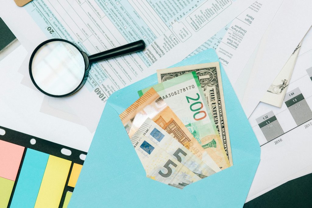 Budget mensuel : comment en créer un facilement et le respecter Close-up of tax documents and mixed currency in a blue envelope with a magnifying glass, perfect for financial analysis concepts.