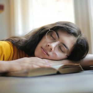 A young woman with glasses naps on a book indoors during the day, capturing a moment of relaxation and fatigue.