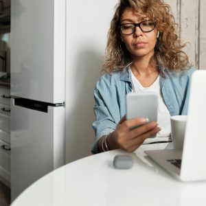 Woman using smartphone and laptop while working remotely in a home kitchen setting.